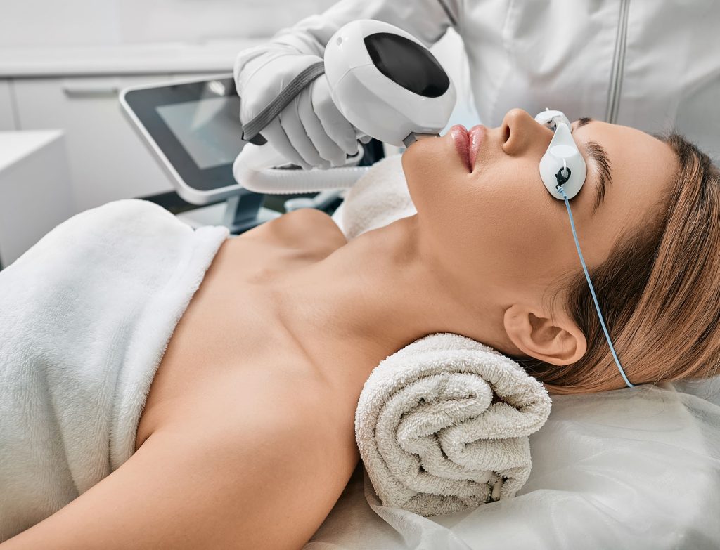 woman receiving facial laser treatment woman lying on a treatment bed with eye protection while a professional uses a handheld IPL device on her face