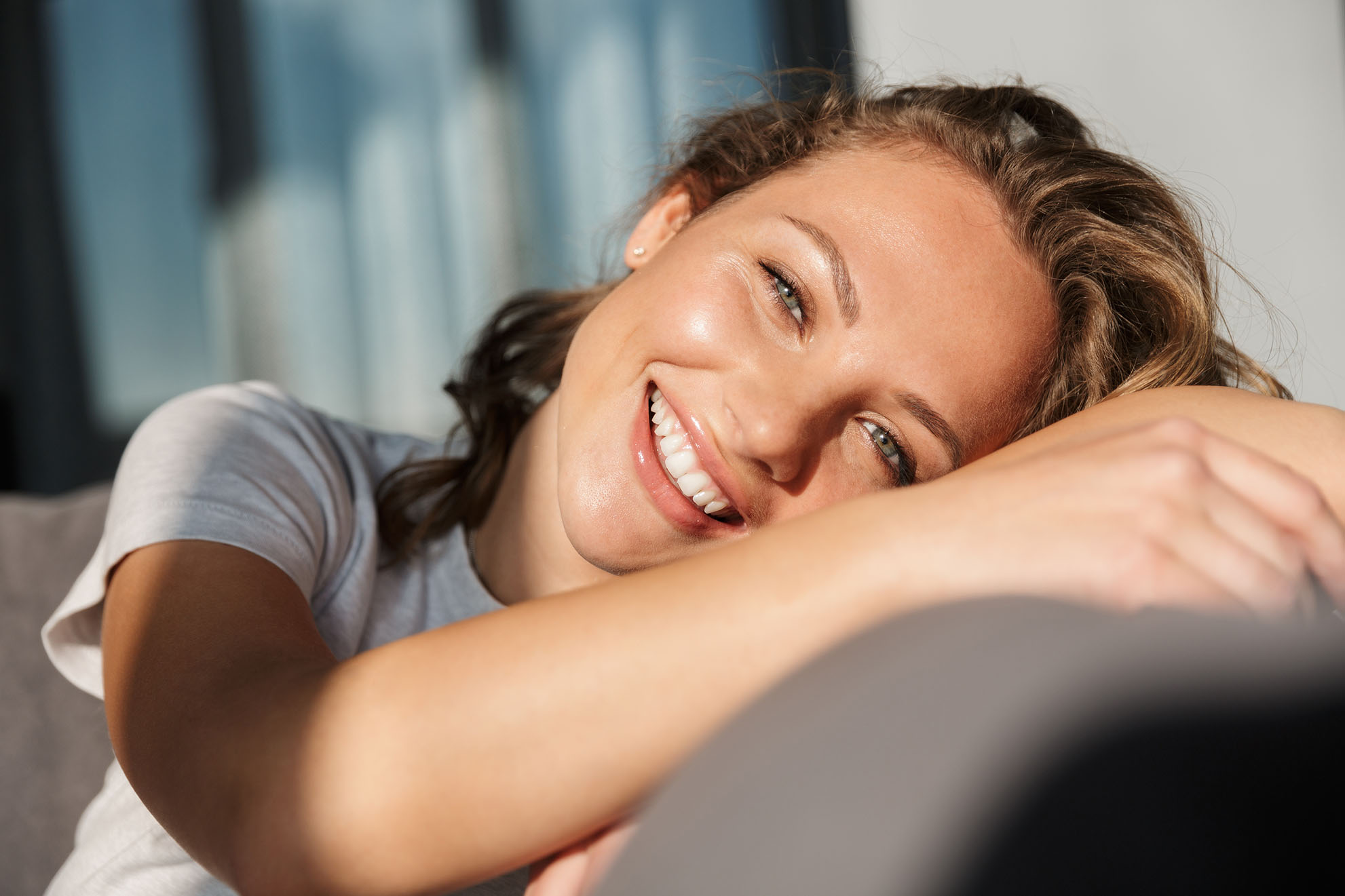 woman with light brown hair leaning on her arm and smiling while sitting in soft sunlight indoors
