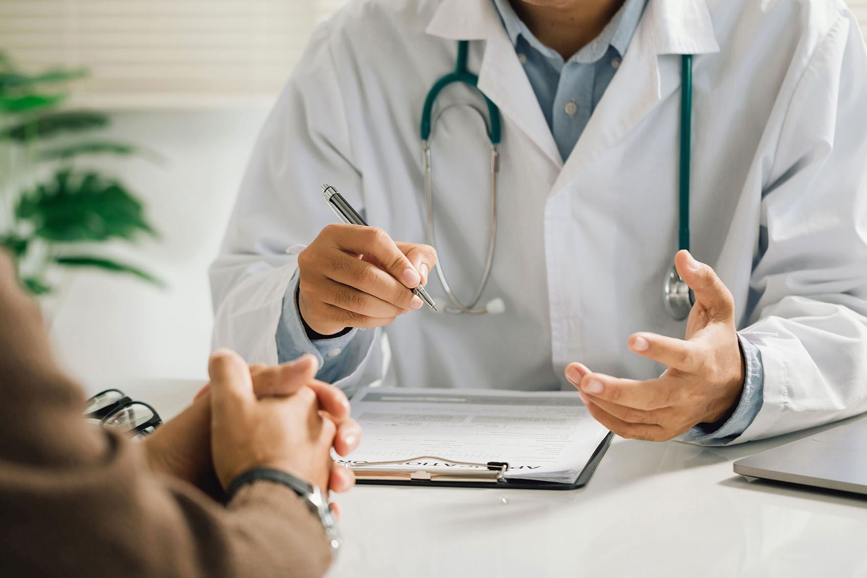 A doctor holding a pen speaks with a patient across a desk with forms and a stethoscope visible.