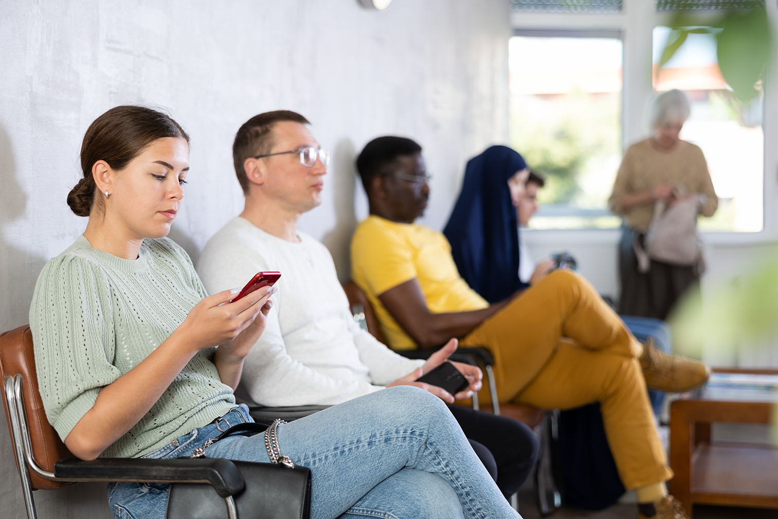 Several people sit in chairs in a waiting room, some using their phones while waiting.