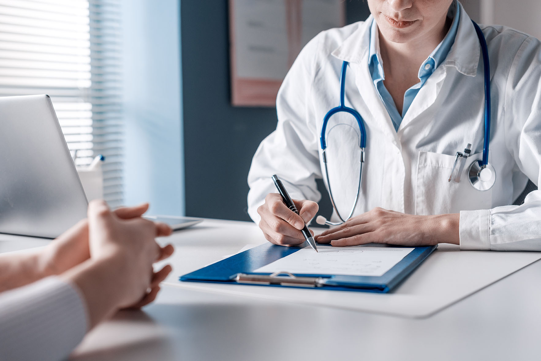 A doctor writes on a clipboard at a desk while a patient sits across with hands folded.