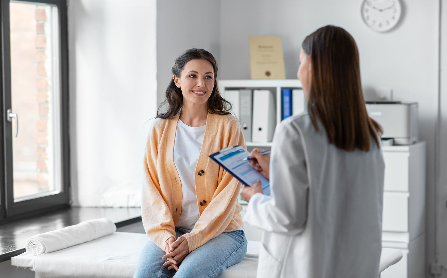 A woman sits on an exam table while a doctor holding a clipboard speaks to her in a bright medical office.