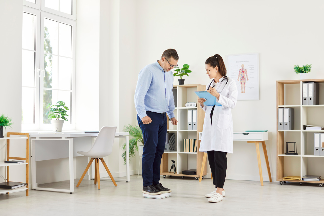 Man stands on a scale while a clinician reviews information on a clipboard nearby